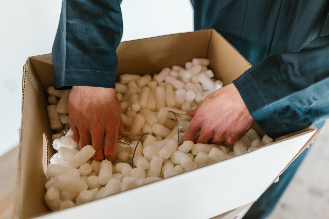 Person carefully packing fragile glassware in a shipping box with polystyrene for protection.